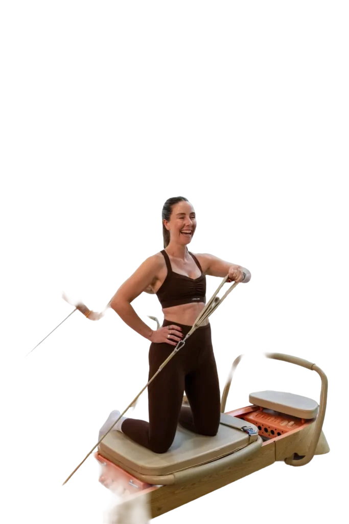 A member kneeling on the reformer, pulling a strap across her body and laughing